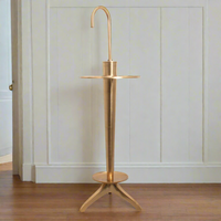 Antiqued brass umbrella stand with a vintage feel, featuring a circular tray on top and a hook at the top for hanging umbrellas. The stand is placed against a white wall on a wooden floor.