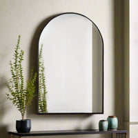 A black thin framed arched mirror mounted on a wall above a console table, with a plant and decorative items to the side.