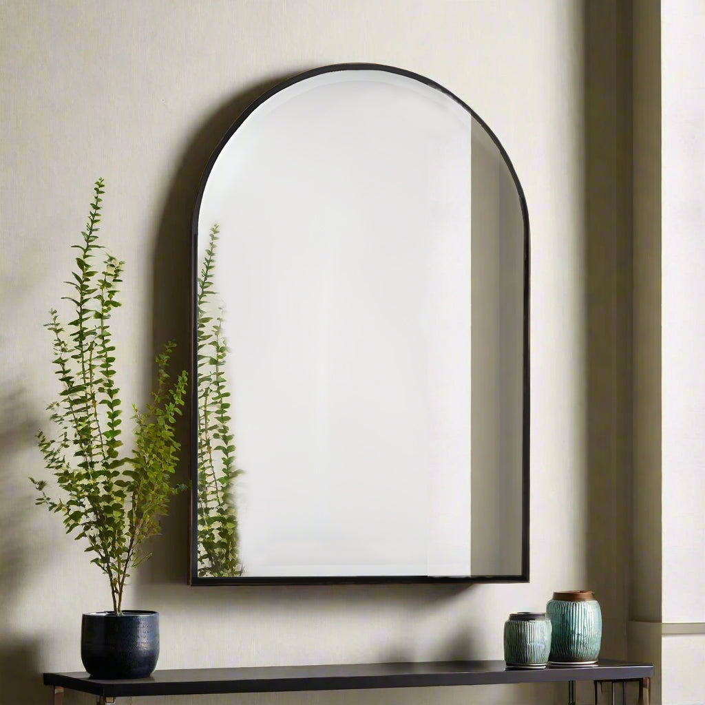 A black thin framed arched mirror mounted on a wall above a console table, with a plant and decorative items to the side.