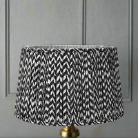 A black pleated lampshade with a chevron pattern, placed on a gold-colored base, with a wall in the background.