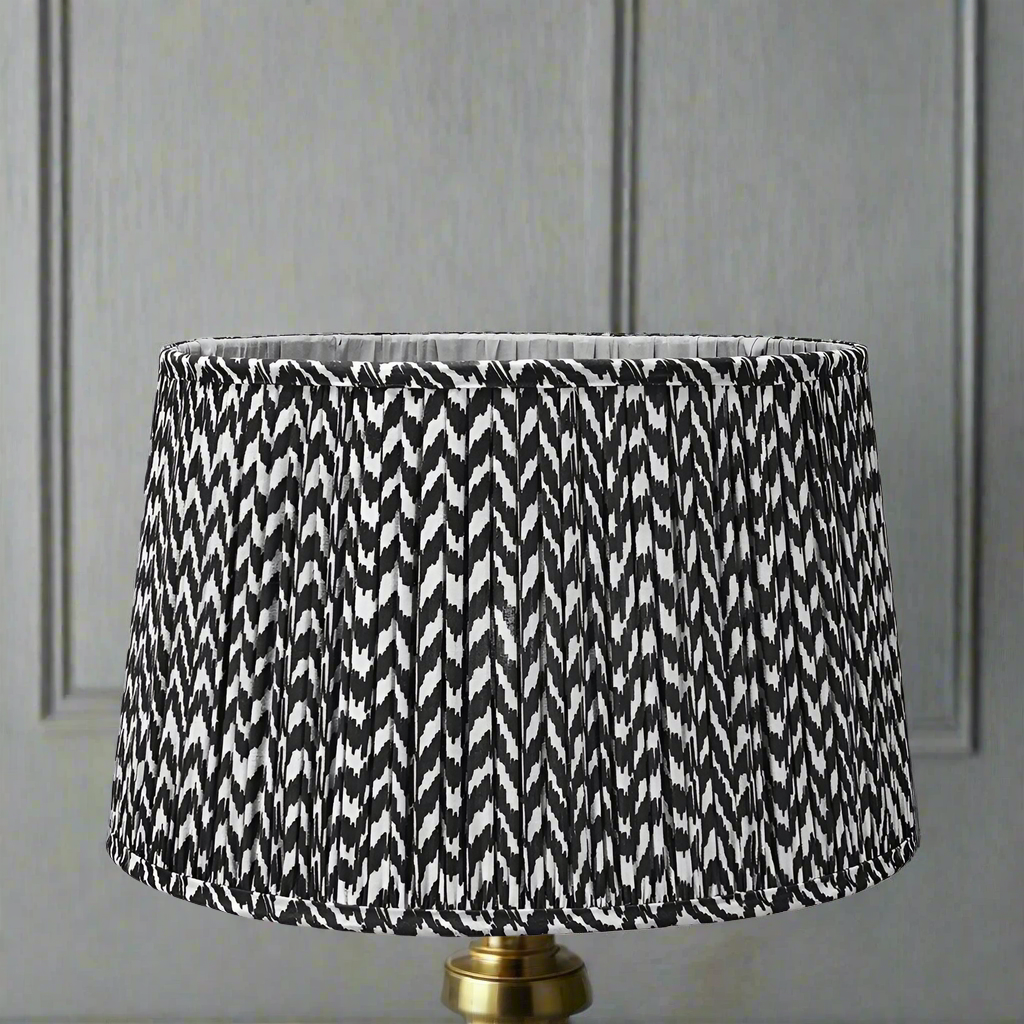 A black pleated lampshade with a chevron pattern, placed on a gold-colored base, with a wall in the background.