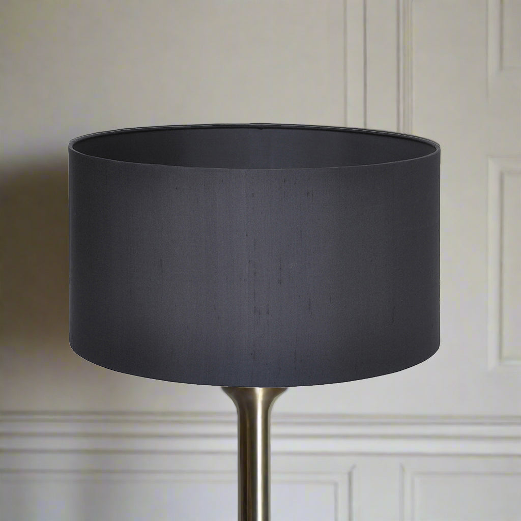 A black silk drum lampshade on a stand, against a wall with a light background.