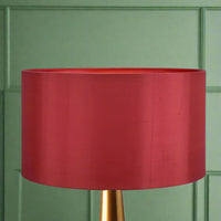 A red silk lampshade with a drum shape, positioned on a stand against a green background.