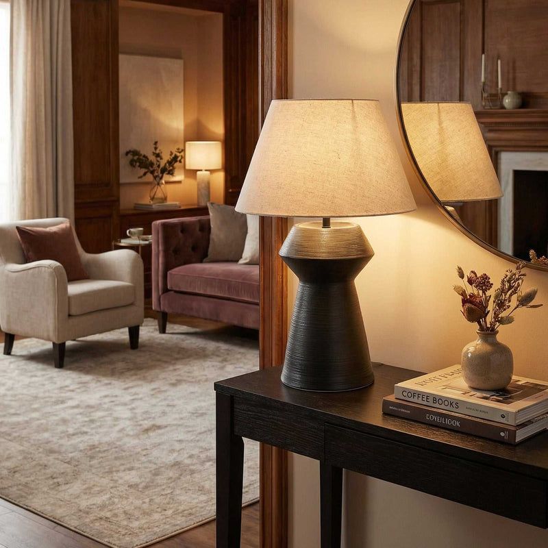 The sculptural black base features a subtle textured finish, beautifully contrasted by a soft neutral fabric shade that diffuses light gently throughout the room. Its balanced proportions make it a versatile piece that complements both modern and classic interiors.