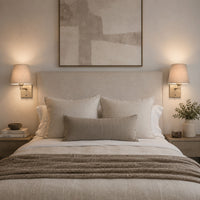 Bedroom with beige upholstered headboard, neutral linen pillows, and warm wall-mounted brass wall lights with taupe shades for cozy lighting
