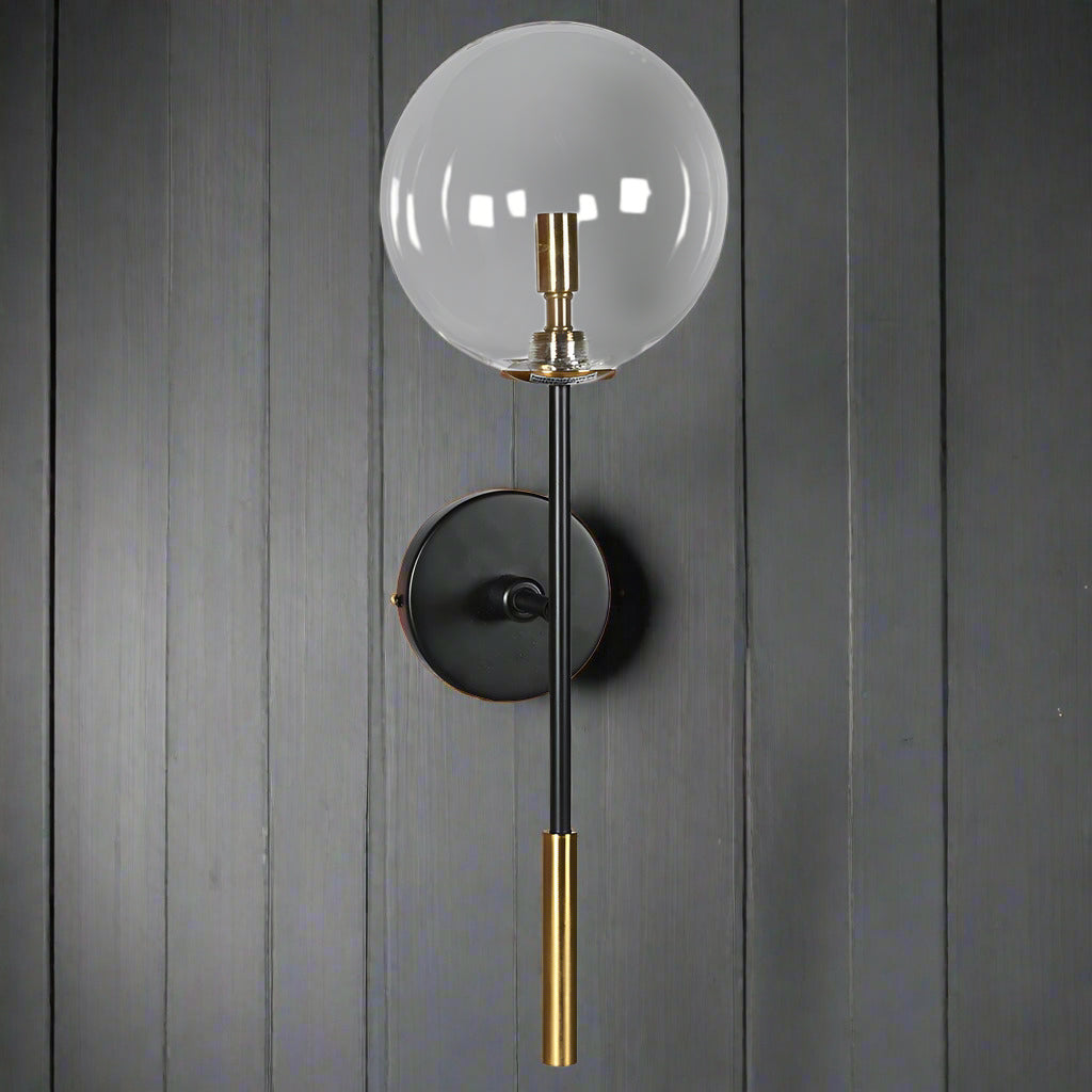 Clear globe wall light set on a circular matte black finish back plate, a long vertical rod extends from the base with a brushed brass cylindrical accent making for a an elegant contrast. Near the top of the rod is a brass socket for the bulb.

The clean lines and black gold combination on this long clear globe wall light evokes a sophisticated aesthetic.&nbsp;&nbsp;

H: 45 cm W: 20 cm D: 25 cm&nbsp;