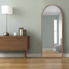 An arched mirror with a natural oak frame, placed against a wall reflecting in the mirror, with a wooden sideboard and lamp to the side.