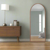 An arched mirror with a natural oak frame, placed against a wall reflecting in the mirror, with a wooden sideboard and lamp to the side.
