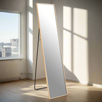 A tall slim framed gold cheval mirror with a stand, placed against a wall in a room with natural lighting.