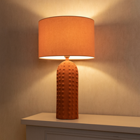 This is a ceramic table lamp finished in a warm terracotta color, featuring a monochromatic design where both the base and the drum-shaped fabric shade share the same earthy hue. The lamp base stands out with its raised bobble texture, giving it a tactile and contemporary appeal. The base is tall and cylindrical with a rounded top, and the bobbled surface adds visual depth and a handcrafted feel, while the uniform terracotta finish provides a cohesive, minimalist look. This lamp is ideal for interiors with 