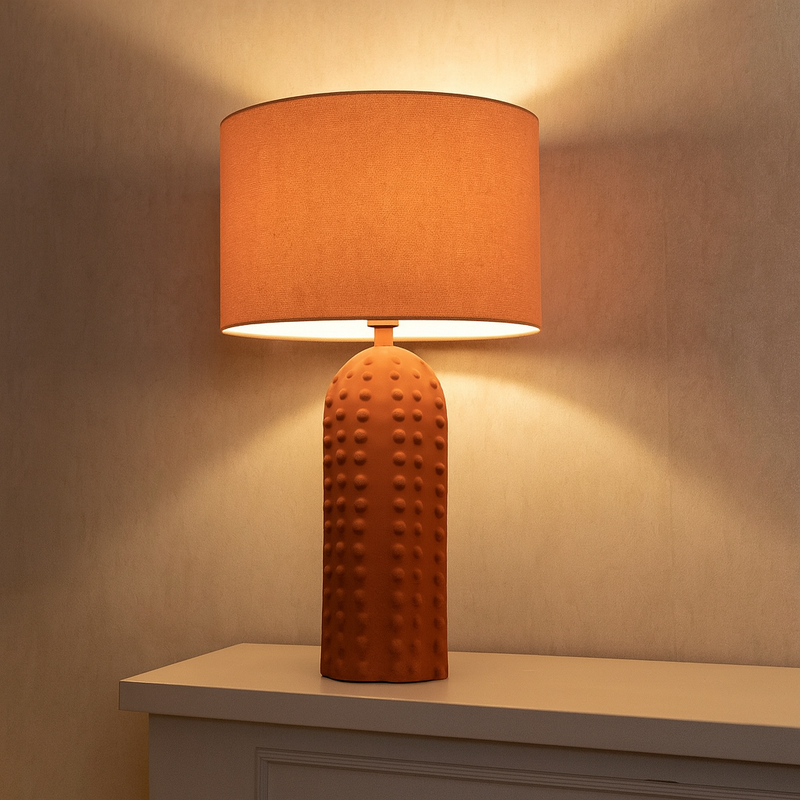 This is a ceramic table lamp finished in a warm terracotta color, featuring a monochromatic design where both the base and the drum-shaped fabric shade share the same earthy hue. The lamp base stands out with its raised bobble texture, giving it a tactile and contemporary appeal. The base is tall and cylindrical with a rounded top, and the bobbled surface adds visual depth and a handcrafted feel, while the uniform terracotta finish provides a cohesive, minimalist look. This lamp is ideal for interiors with 
