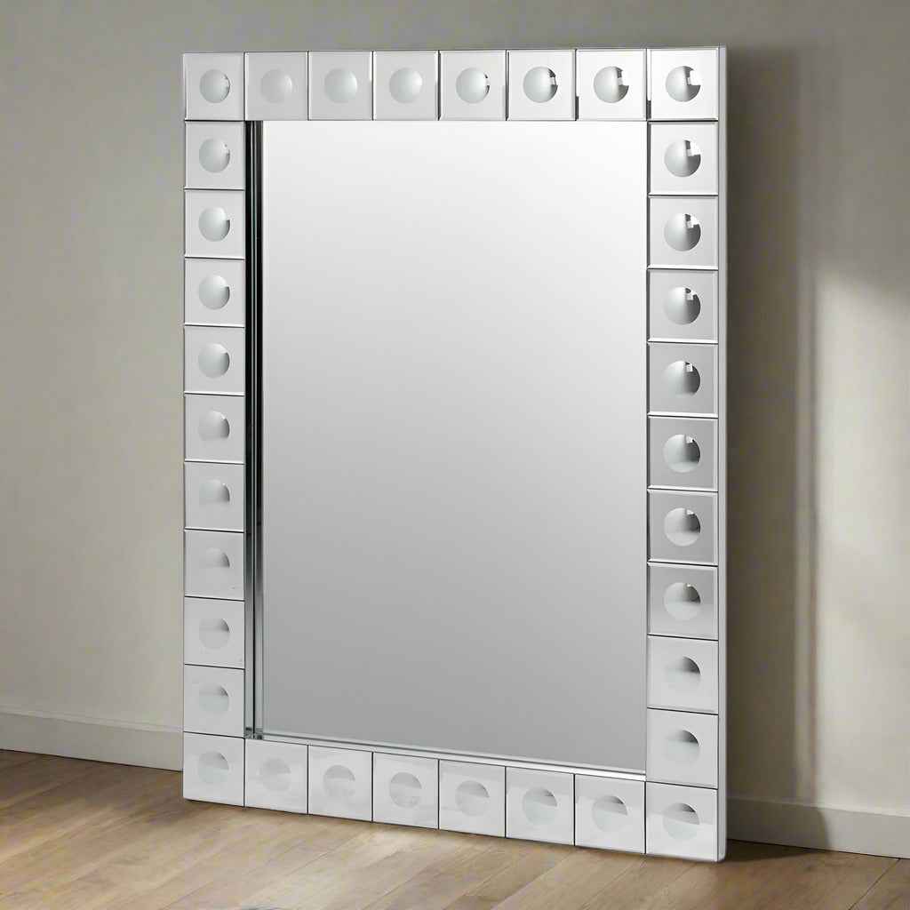 This is a Venetian-style wall mirror featuring a striking raised decorative frame. The rectangular mirror is bordered by a geometric design of square mirrored tiles, each accented with a raised circular dome detail that adds dimension, texture, and light-catching elegance. The reflective frame enhances the mirror’s brilliance, making it not only functional but also a bold decorative statement.

Its clean lines and sculptural frame give it a contemporary twist on classic Venetian design, making it versatile 