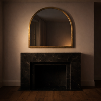 Brass overmantle over a black marble fireplace in a minimalist room with moody lighting and bare wooden floor.