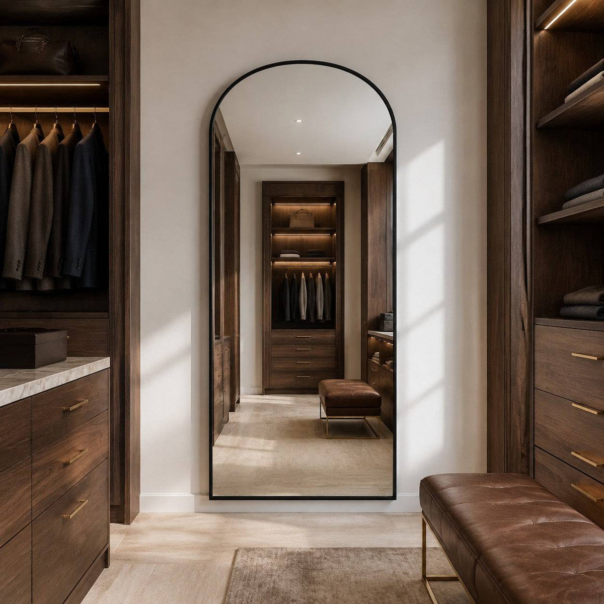 A tall arched floor mirror with a thin black frame stands in a walk-in closet, reflecting a matching wood storage area. The room features dark wood cabinetry, gold-tone handles, a brown leather bench, and warm lighting.