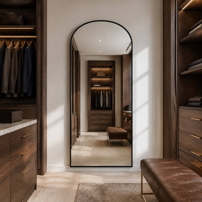 A tall arched floor mirror with a thin black frame stands in a walk-in closet, reflecting a matching wood storage area. The room features dark wood cabinetry, gold-tone handles, a brown leather bench, and warm lighting.