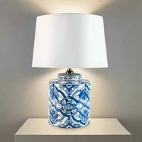 Blue and white lamp, lit on a pale wooden stand against a similar coloured wall,


