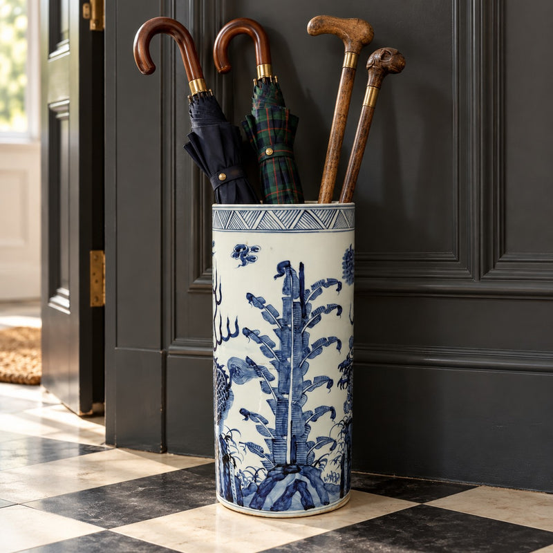 A tall cylindrical ceramic umbrella stand with a blue and white floral pattern, holding several umbrellas and walking canes in front of a dark paneled door.