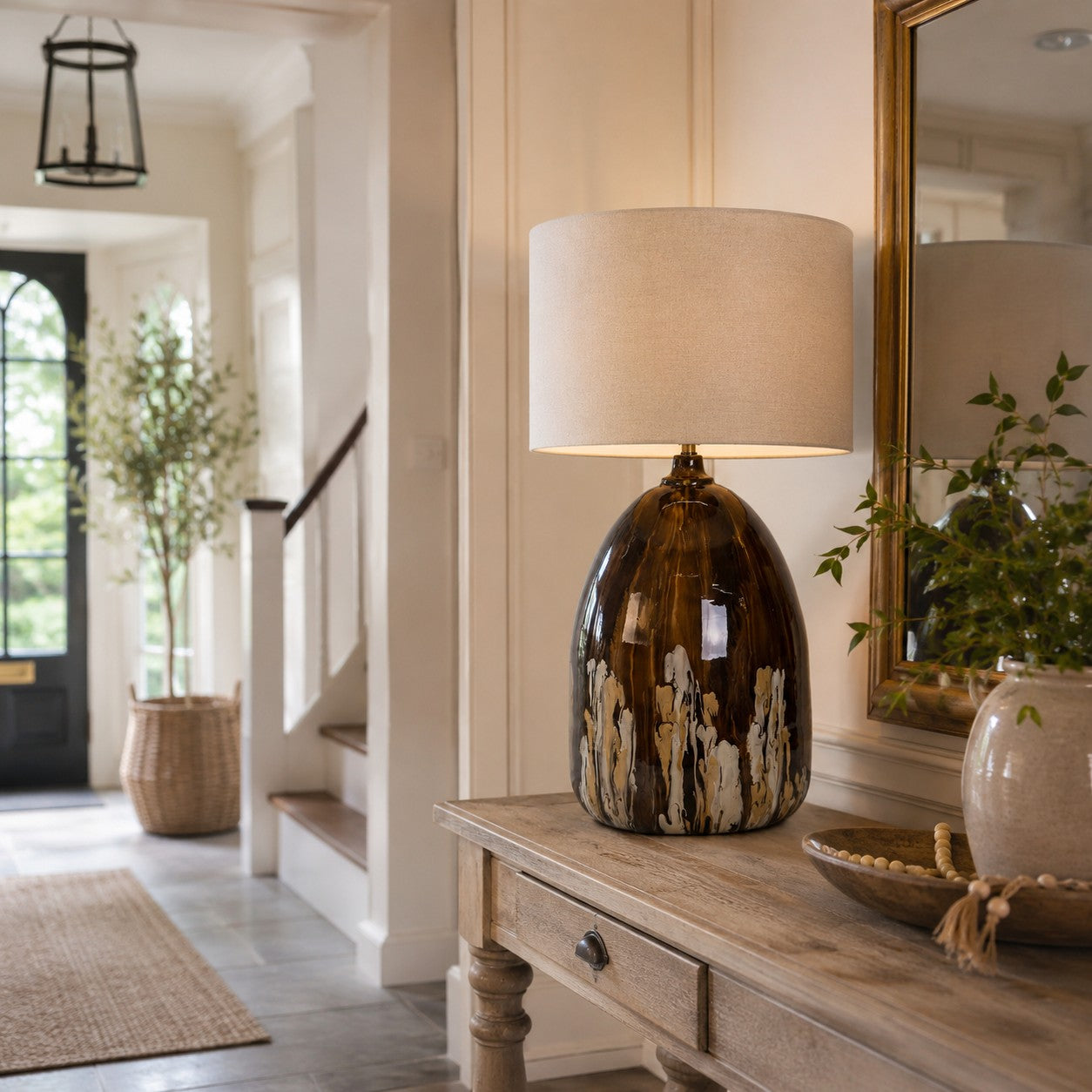 A table lamp with a glossy amber-brown ceramic base and a beige fabric drum shade, styled on a rustic wooden console table in a bright, elegant entryway.