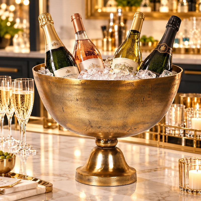 A large gold pedestal bowl filled with ice and four champagne bottles, set on a marble countertop in a warmly lit bar or lounge setting.
