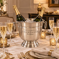 A silver champagne bucket filled with ice and two champagne bottles,  on a decorated dining table with champagne flutes, gold candle holders, in a warm elegant room.