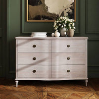 Soft off white six drawer chest with round brass knobs, wooden furniture piece for bedroom or living room storage, against dark green paneled wall