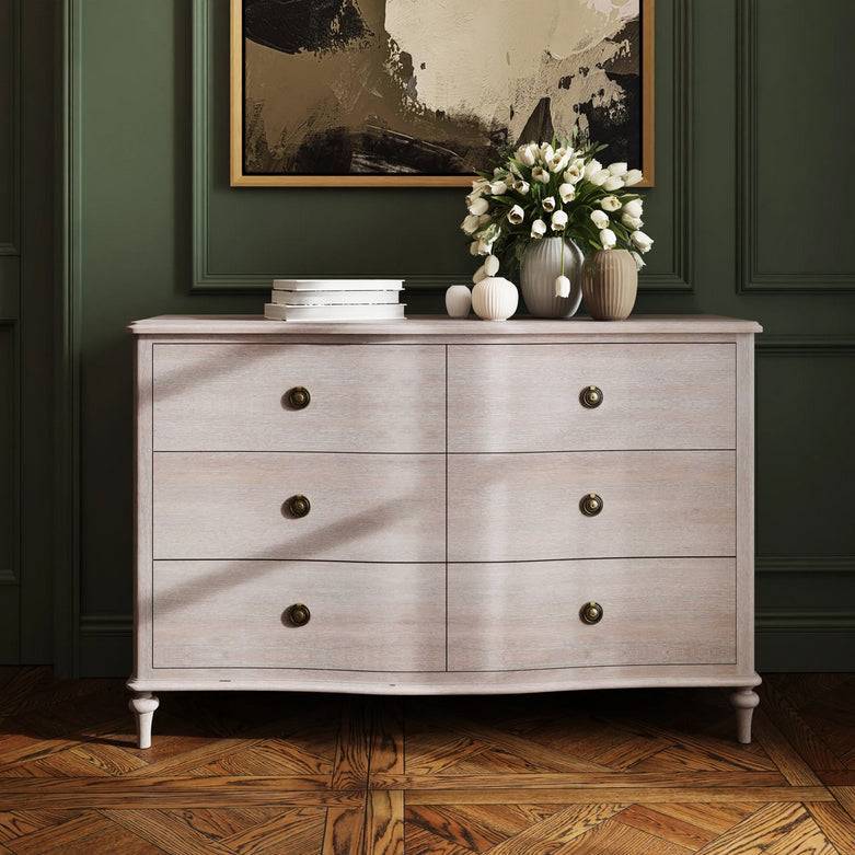 Soft off white six drawer chest with round brass knobs, wooden furniture piece for bedroom or living room storage, against dark green paneled wall
