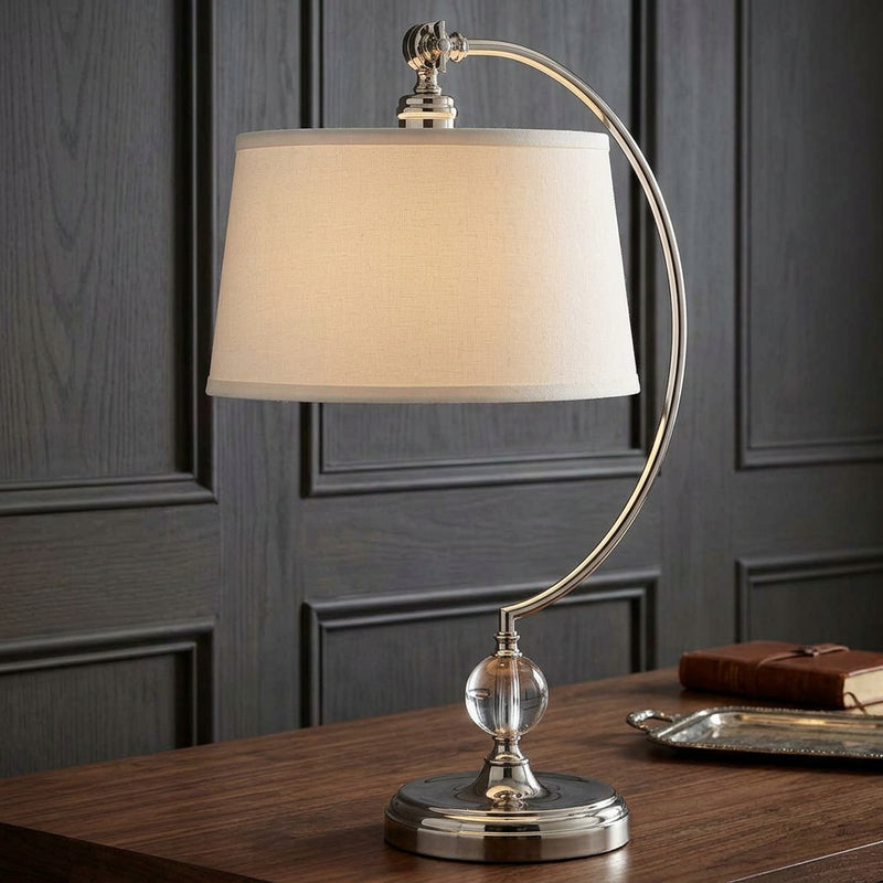 A modern table lamp with a curved silver metal arc, a clear glass sphere accent, and a tapered off-white fabric shade on a hall table with grey background.