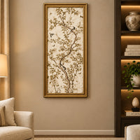 A tall framed wall art piece with a gold border. The artwork shows delicate tree branches with leaves and small birds in soft beige, brown.