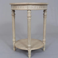 A vintage-inspired limewashed round wooden side table with turned fluted legs and a lower shelf, photographed against a neutral backdrop.