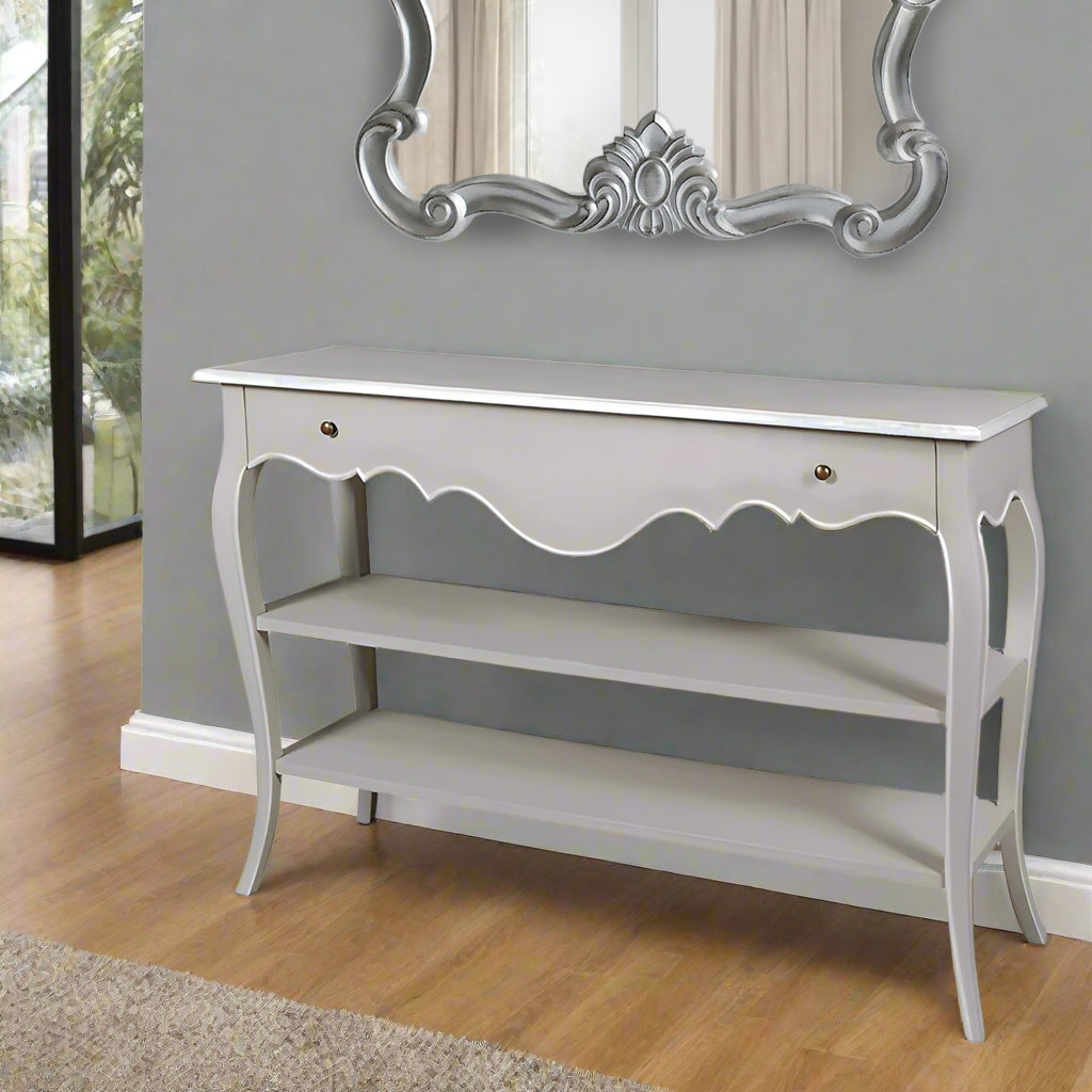 A light grey hand-painted console table with curvy legs, featuring a drawer and two shelves, placed against a wall in a room with a large mirror on the opposite wall.