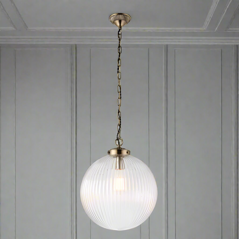 Antique brass and clear ribbed glass globe pendant light suspended from a decorative chain, showcasing a warm glowing bulb inside.