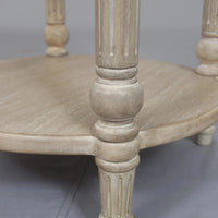 A vintage-inspired limewashed round wooden side table with turned fluted legs and a lower shelf, photographed against a neutral backdrop.