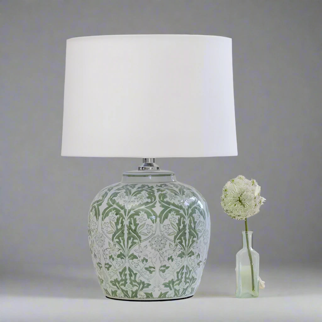 Decorative table lamp with green floral pattern next to a small vase with flowers on a gray background