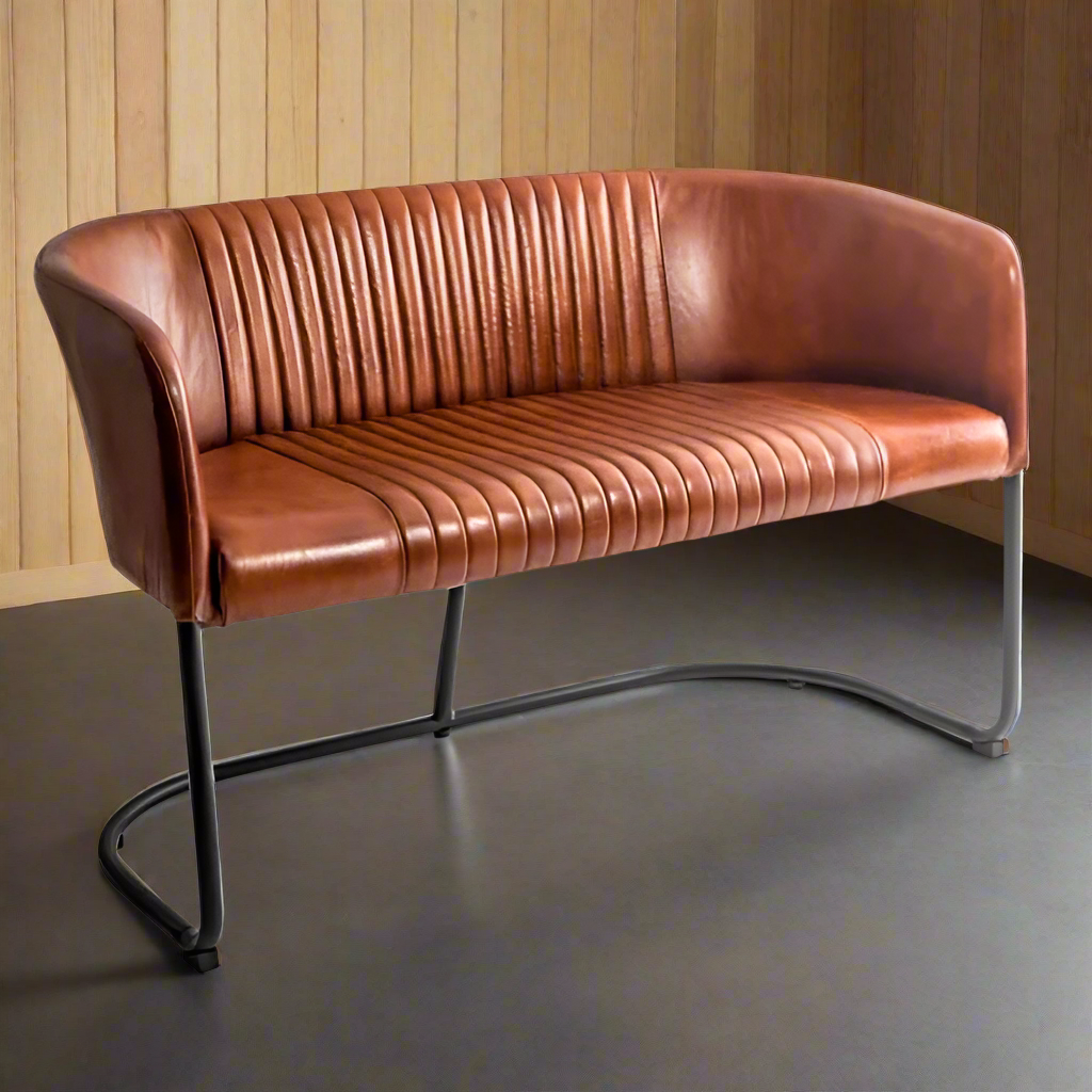 A brown vintage leather two seat sofa with black iron legs, positioned against a wooden wall on a grey floor.