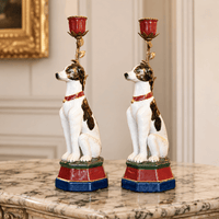 Pair of seated white porcelain dog candleholders with brown markings, red collars, colourful stepped bases and ornate floral stems holding red candle cups.