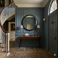 The oversize black wooden mirror is shown in a traditional dark blue hallway next to  a sweeping staircase , over a wooden console with brass lighting. 