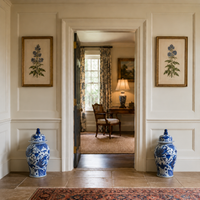 A symmetrical interior hallway with two blue-and-white porcelain lidded vases on the floor, framed botanical prints on the walls, and a doorway leading into a warmly lit study with floral curtains and a desk lamp.
