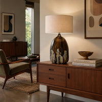 A mid-century modern table lamp with a glossy brown and gold marbled base and a beige fabric drum shade, styled on a wooden credenza in a warm, contemporary living room.