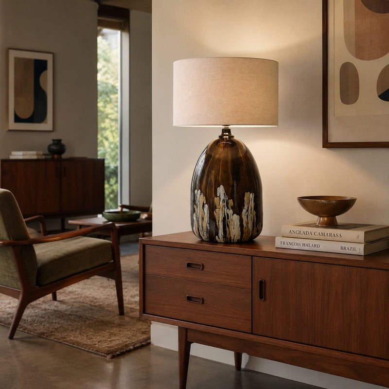 A mid-century modern table lamp with a glossy brown and gold marbled base and a beige fabric drum shade, styled on a wooden credenza in a warm, contemporary living room.