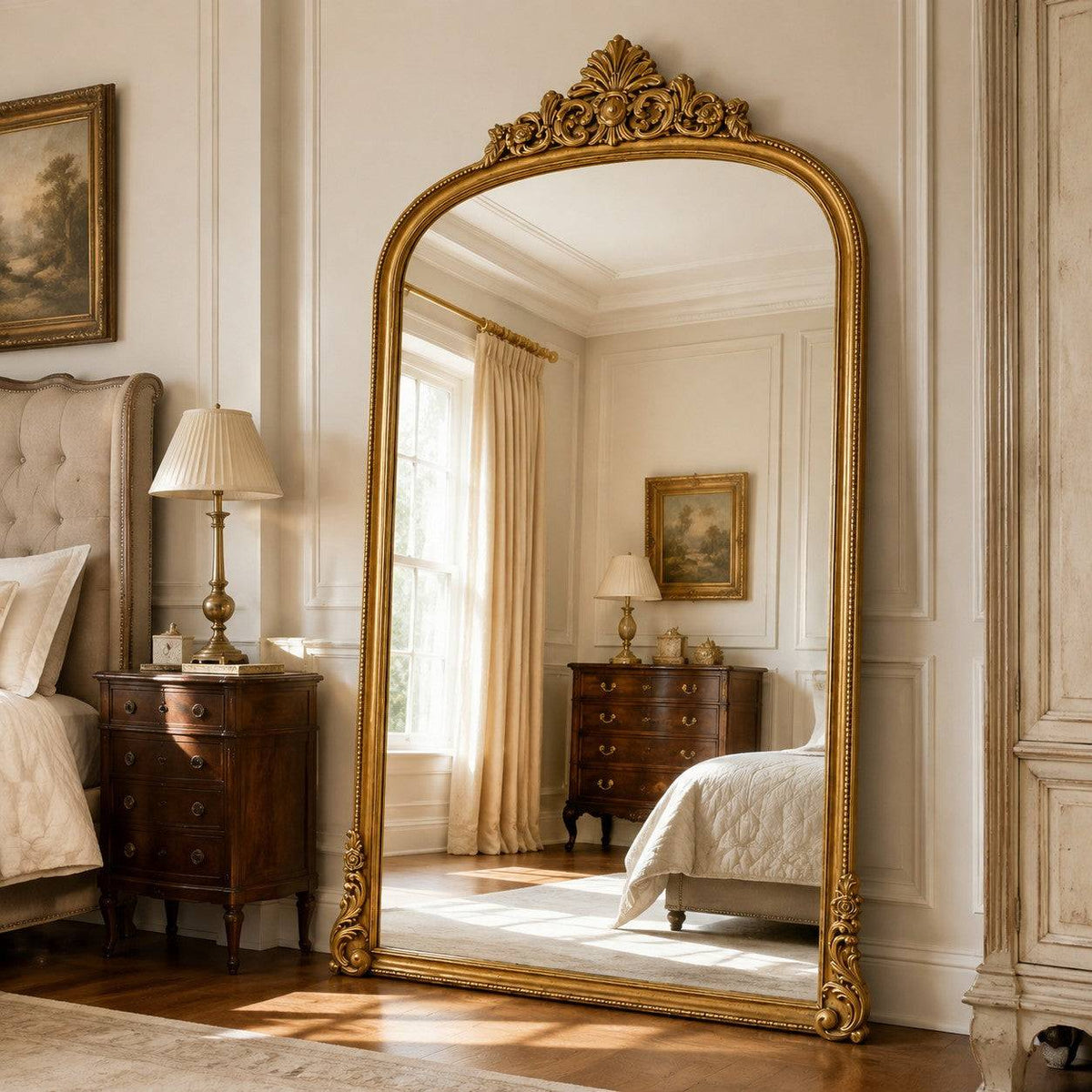 Gilt arched leaner mirror with ornate gold frame and decorative crest, reflecting a classic bedroom with vintage furniture and soft lighting