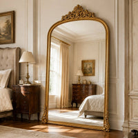 Gilt arched leaner mirror with ornate gold frame and decorative crest, reflecting a classic bedroom with vintage furniture and soft lighting