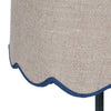 A slim very dark green, metal table lamp with a traditional design. It features a slender, ornate stand and a beige linen lampshade with a scalloped edge trimmed in blue. 