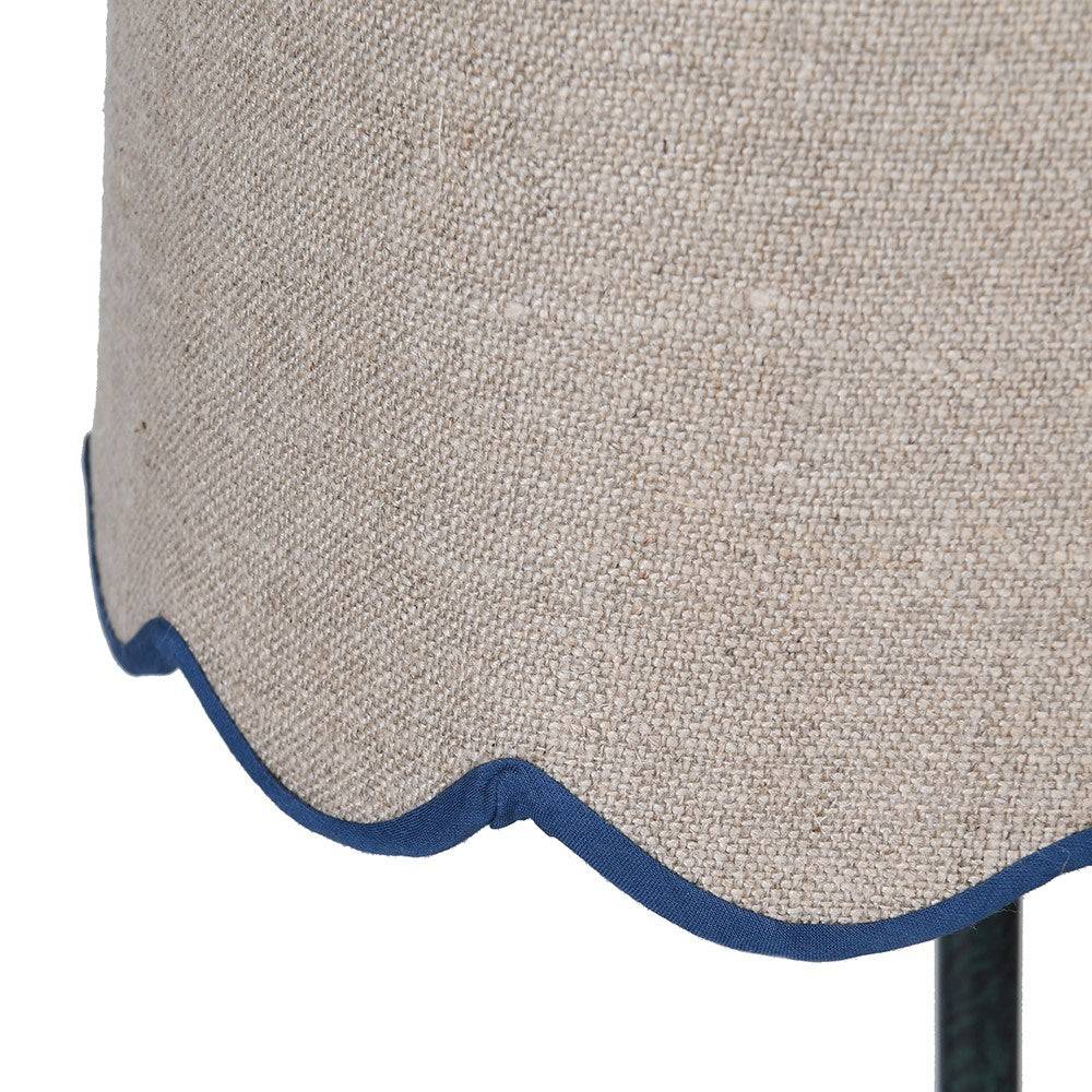 A slim very dark green, metal table lamp with a traditional design. It features a slender, ornate stand and a beige linen lampshade with a scalloped edge trimmed in blue. 