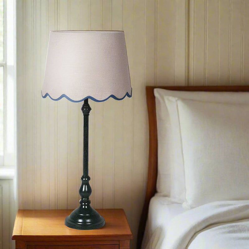 A slim very dark green, metal table lamp with a traditional design. It features a slender, ornate stand and a beige linen lampshade with a scalloped edge trimmed in blue. 