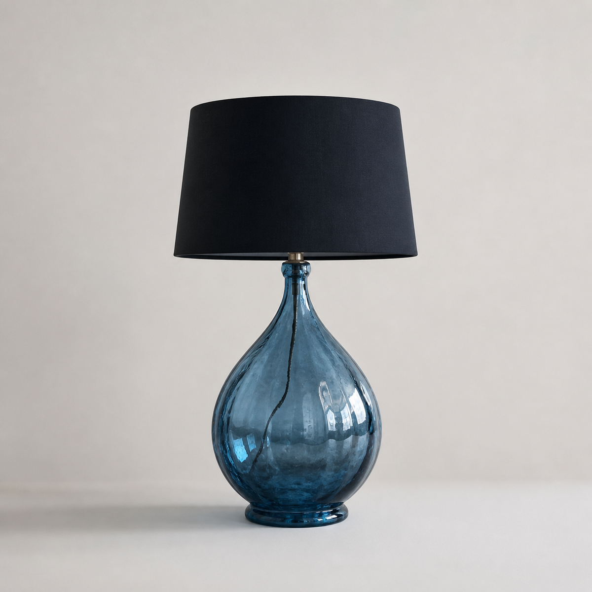 Oversized blue glass table lamp with teardrop-shaped base and black drum shade on neutral background, modern statement lighting piece.