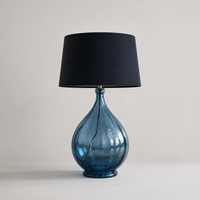 Oversized blue glass table lamp with teardrop-shaped base and black drum shade on neutral background, modern statement lighting piece.