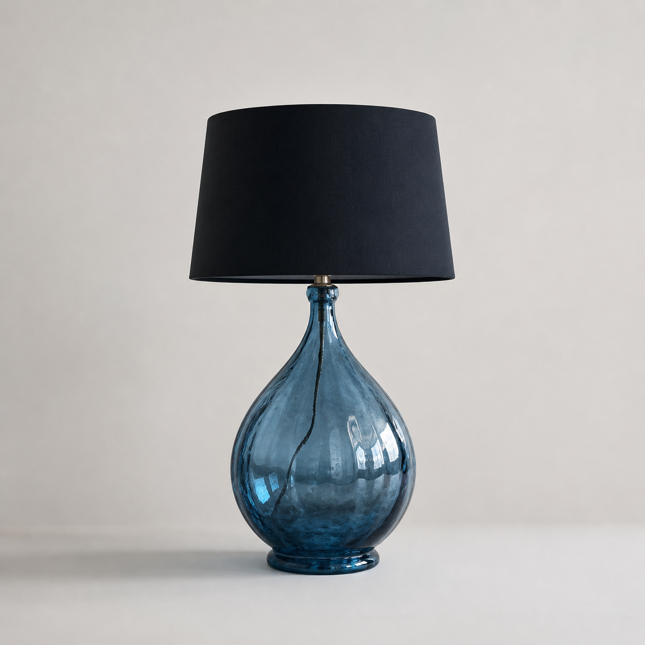 Oversized blue glass table lamp with teardrop-shaped base and black drum shade on neutral background, modern statement lighting piece.