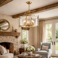 Gold lantern chandelier with clear glass drum shade hanging in a rustic farmhouse living room with exposed wood beams and stone fireplace.