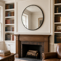 A round black-framed mirror with a distressed textured edge hangs on a cream wall above a dark wood fireplace. The room features built-in bookshelves filled with books, a leather armchair in the foreground, and a window on the left.