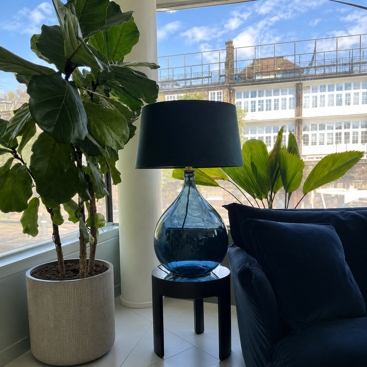 Blue lamp on a small table with a plant and sofa in a room with large windows.